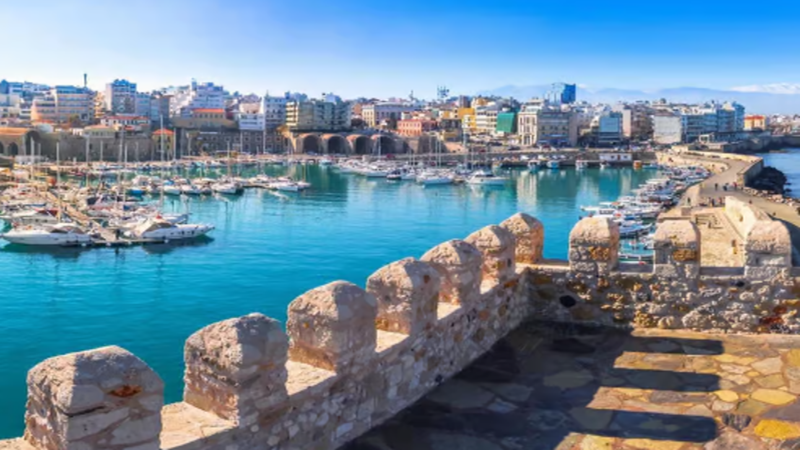 Architectural skyline of Heraklion port and modern commercial infrastructure in Crete with digital blockchain network overlay graphics.