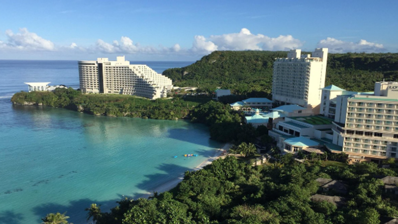 A luxurious tropical architectural rendering of a Tumon Bay oceanfront resort development in Guam integrated with digital blockchain overlay graphics.